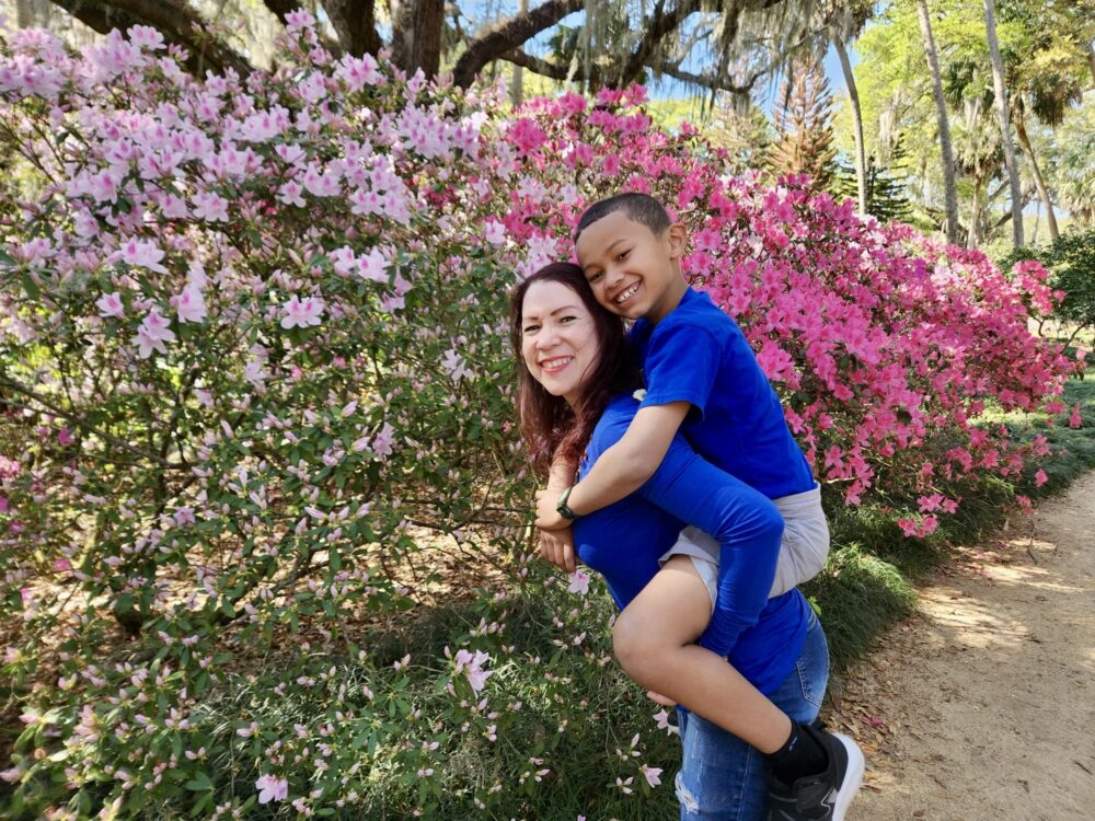 A smiling woman carries a young child on her back while standing in front of pink and white flowering shrubs in a garden. Both are wearing blue shirts and smiling at the camera on a sunny day.