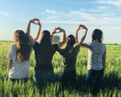 Four friends standing in a green field, forming heart shapes with their hands under a blue sky.