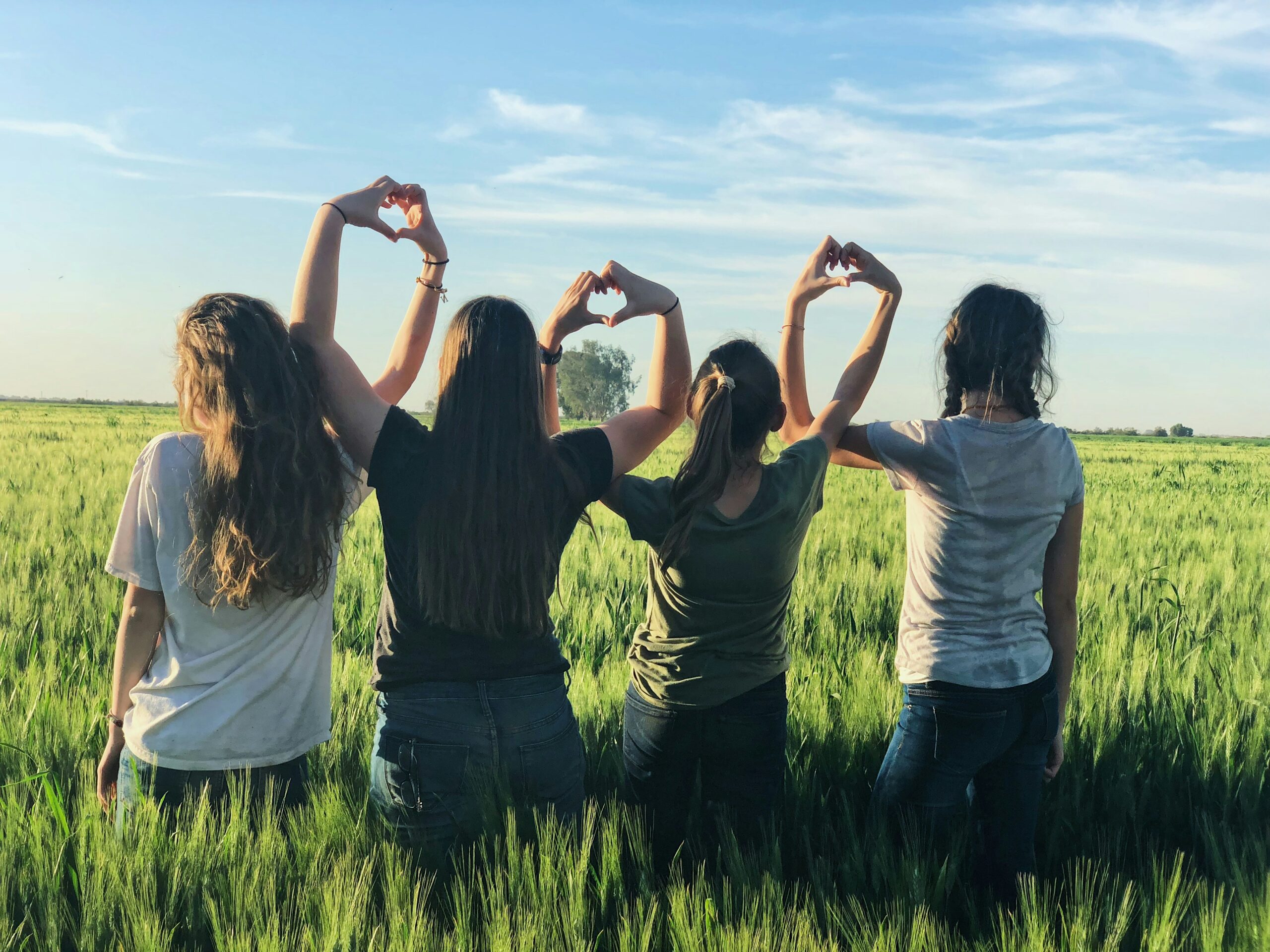 Four friends standing in a green field, forming heart shapes with their hands under a blue sky.
