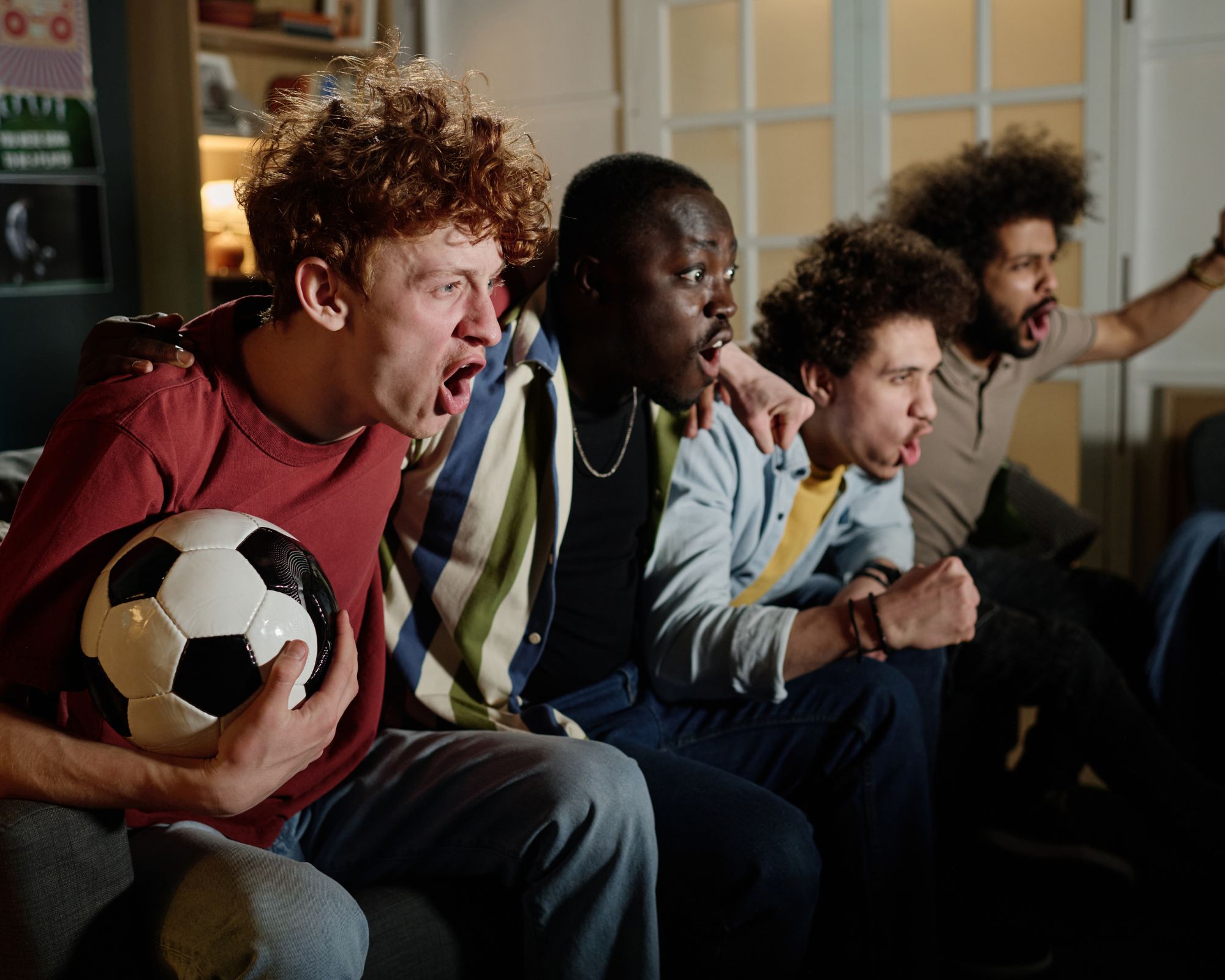 Un grupo de amigos observa un partido de fútbol en televisión mientras reacciona con emoción; uno de ellos sostiene un balón de fútbol y todos miran atentamente hacia la pantalla.