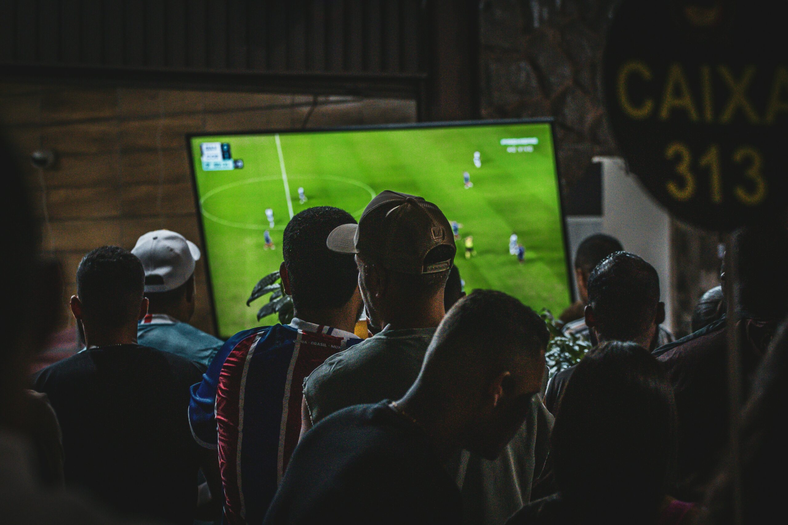 A group of people watch a live soccer match on a large television in a crowded indoor gathering, with fans standing closely together and focused on the game.